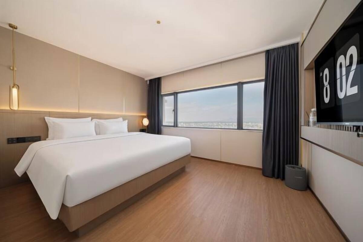 A modern hotel room with a large bed, a view of the city, and a digital clock displaying 8:02.