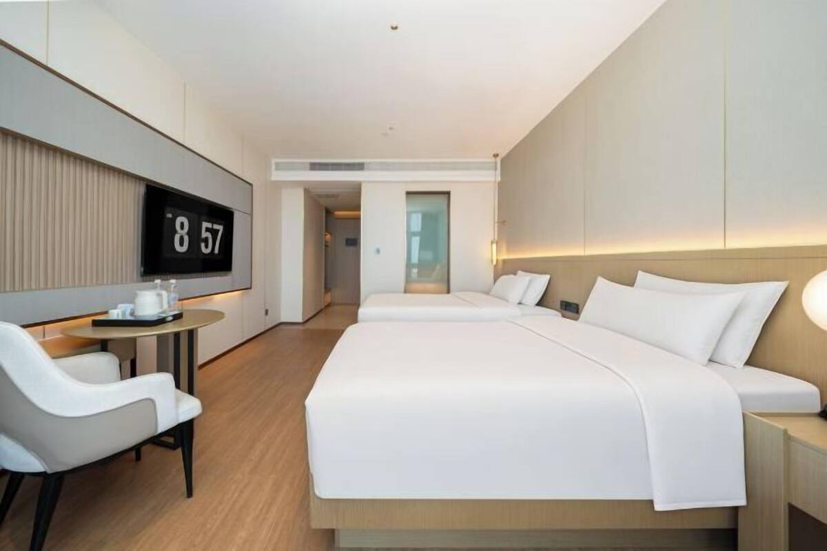 A modern hotel room with a large bed, a bedside table, a chair, a small table with a coffee maker, and a digital clock displaying the time.