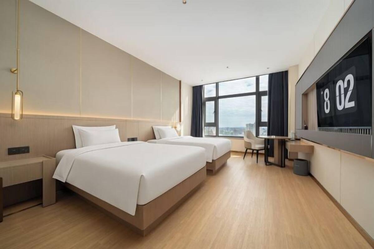 A modern hotel room with two beds, a desk, and a large window offering a city view.