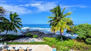 A beach with palm trees, ocean waves, and a patio with lounge chairs and a table. - Luxury Oceanfront Private Estate, large family gatherings (Kailua-Kona)