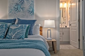 Une chambre à coucher avec un lit, des oreillers, une table de chevet avec une lampe, une œuvre d’art murale et une vue sur la salle de bain.