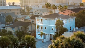 A multi-story residential building with a balcony, surrounded by palm trees and other houses.