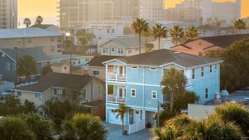 A multi-story residential building with a balcony, surrounded by palm trees and other houses.