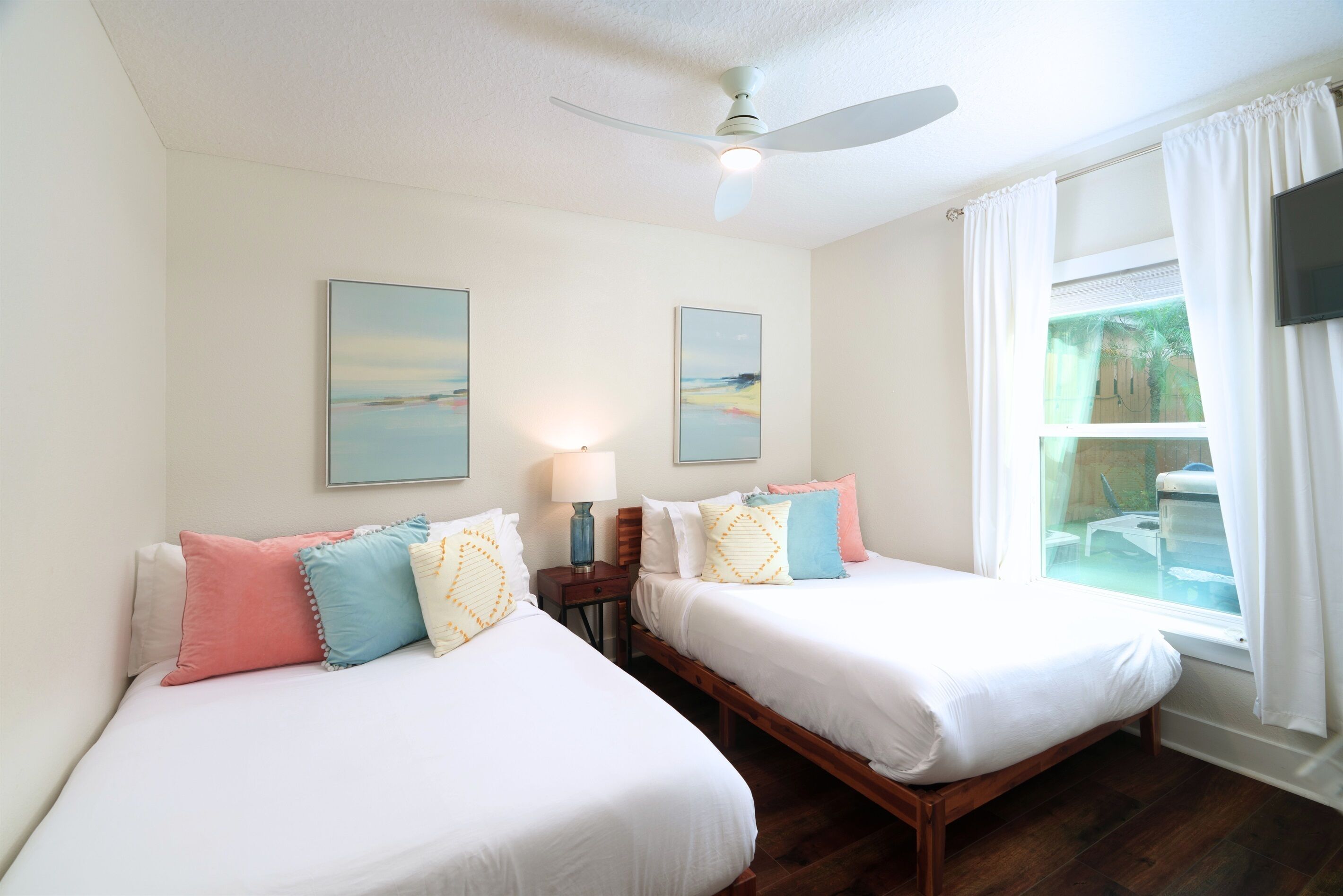 A hotel room with two beds, a ceiling fan, artwork on the walls, and a window with curtains.