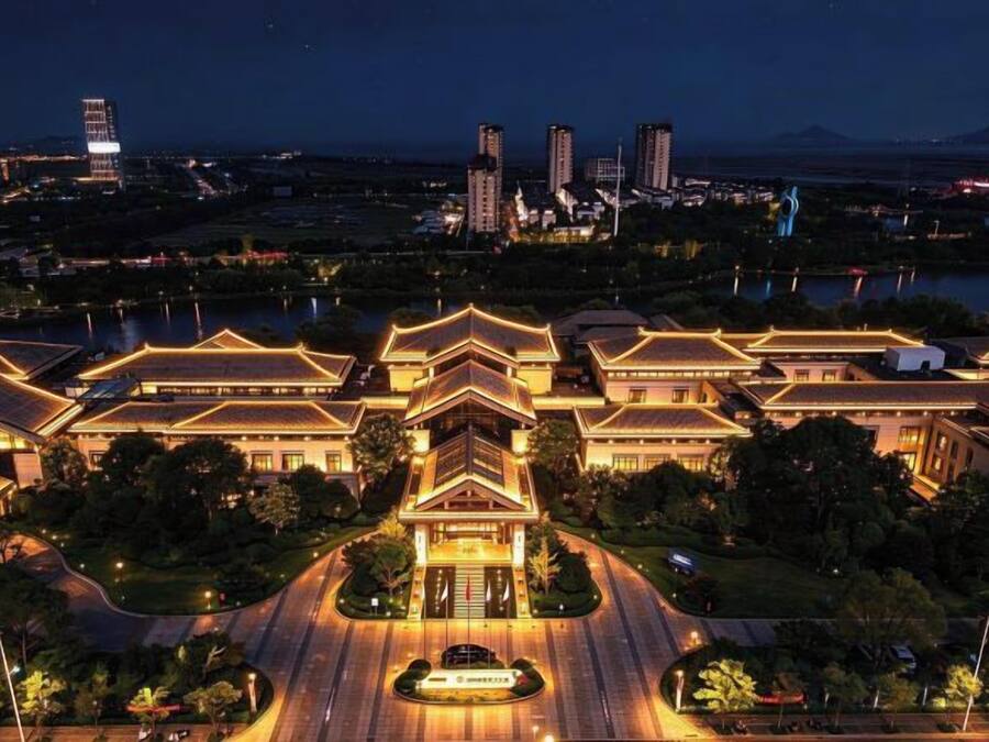 Um edifício tradicional com vários telhados e uma entrada central, rodeado por vegetação e um corpo de água.