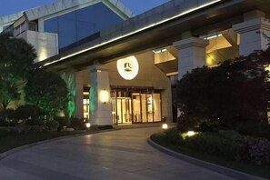 Entrada de un hotel con un gran logotipo iluminado, un vestíbulo techado y un jardín ajardinado.