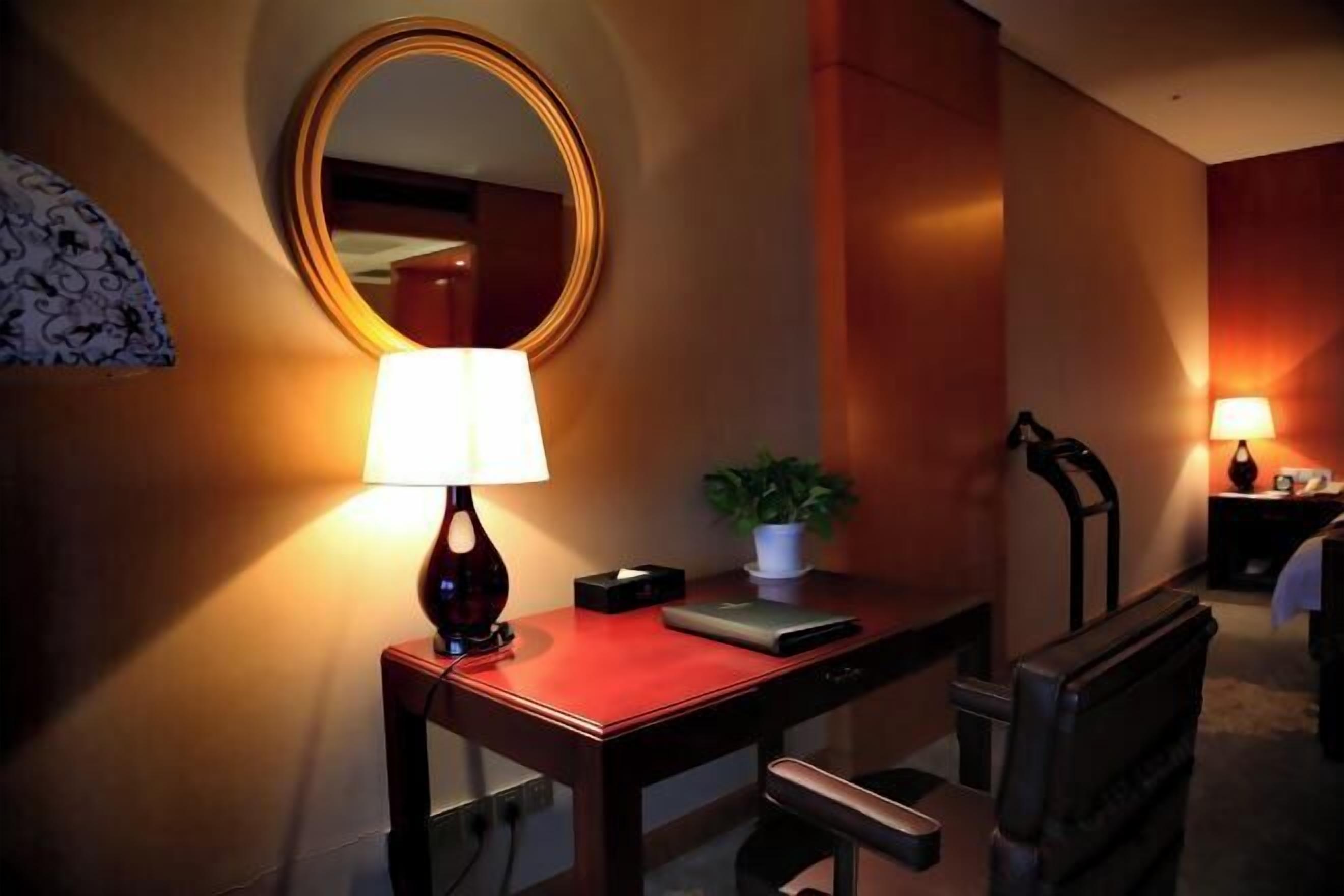A hotel room with a desk, a lamp, a mirror, a chair, and a bedside table with a lamp.