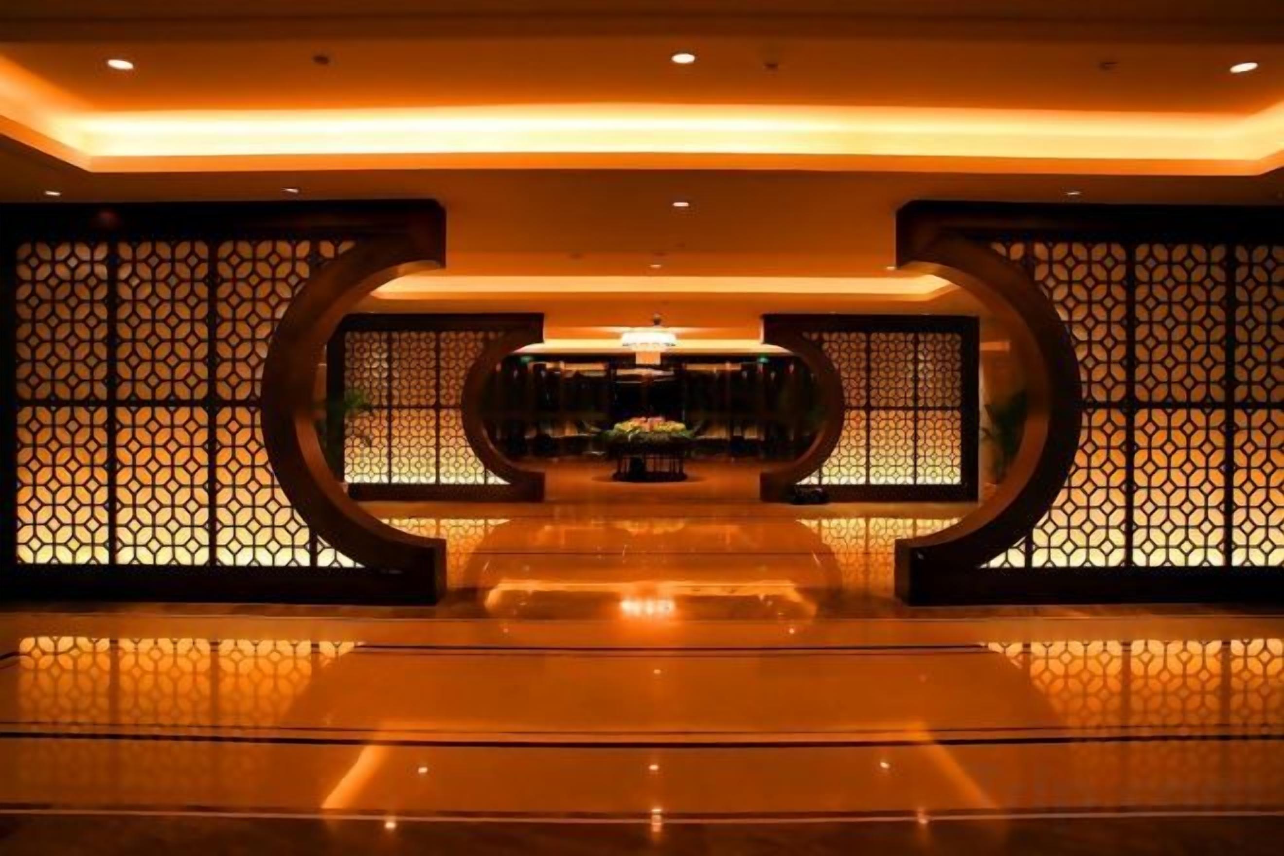 Um hall de hotel com uma mesa de recepção, uma parede decorativa com padrão geométrico e um chão espelhado.