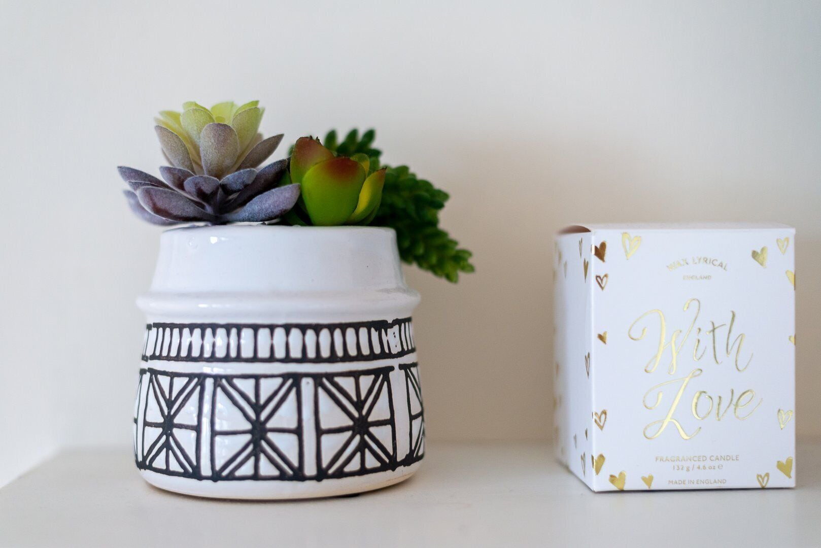 Un pot en céramique blanche orné de motifs géométriques noirs et contenant une plante succulente. À côté, on trouve une boîte blanche contenant une bougie, avec des lettres dorées et des motifs en forme de cœur.