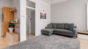 A modern living room with a grey sectional sofa, a grey carpet, and a wooden cabinet with a small shelf. - GuestReady - Beautiful Apt in Clapham (London)