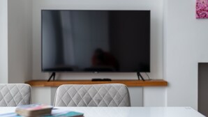 A modern living room with a flat-screen TV mounted on the wall, a white table with a few books, and two upholstered chairs. - GuestReady - Beautiful Apt in Clapham (London)