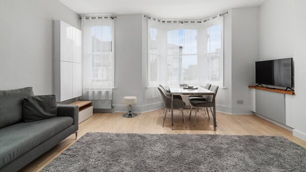 A modern living room with a grey sofa, a dining table with chairs, a small white table lamp, and a flat-screen TV mounted on the wall. - GuestReady - Beautiful Apt in Clapham (London)
