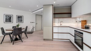 A modern kitchen with white cabinets, a marble countertop, and a built-in oven. There is a dining area with a table and chairs, and two framed pictures on the wall. - GuestReady - 2BR Luxury Flat in Central London (London)