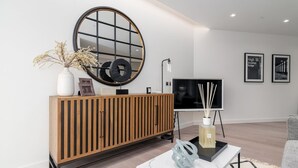 A modern living room with a wooden console, a round mirror, a flat-screen TV, and a coffee table with decorative items. - GuestReady - 2BR Luxury Flat in Central London (London)