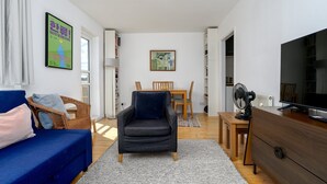A modern living room with a blue sofa, a dark armchair, a wooden dining table with chairs, a TV, and a fan. - GuestReady Essential - Penthouse Vibes in Peckham (London)