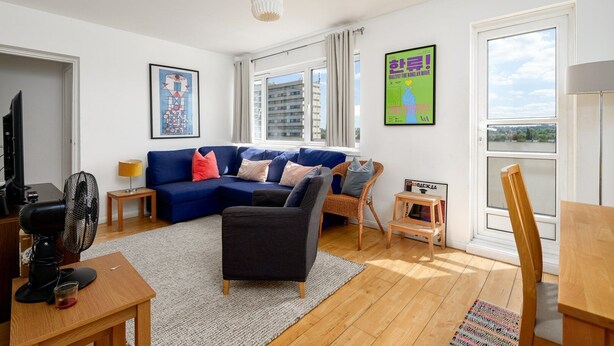 A modern living room with a blue sectional sofa, a small table with a lamp, a TV, and a fan. There are two windows with curtains, a poster on the wall, and a view of buildings outside. - GuestReady Essential - Penthouse Vibes in Peckham (London)
