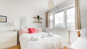 A modern bedroom with a large bed, white bedding, a bedside table, a lamp, a window with curtains, and a view of the city. - GuestReady Essential - Penthouse Vibes in Peckham (London)