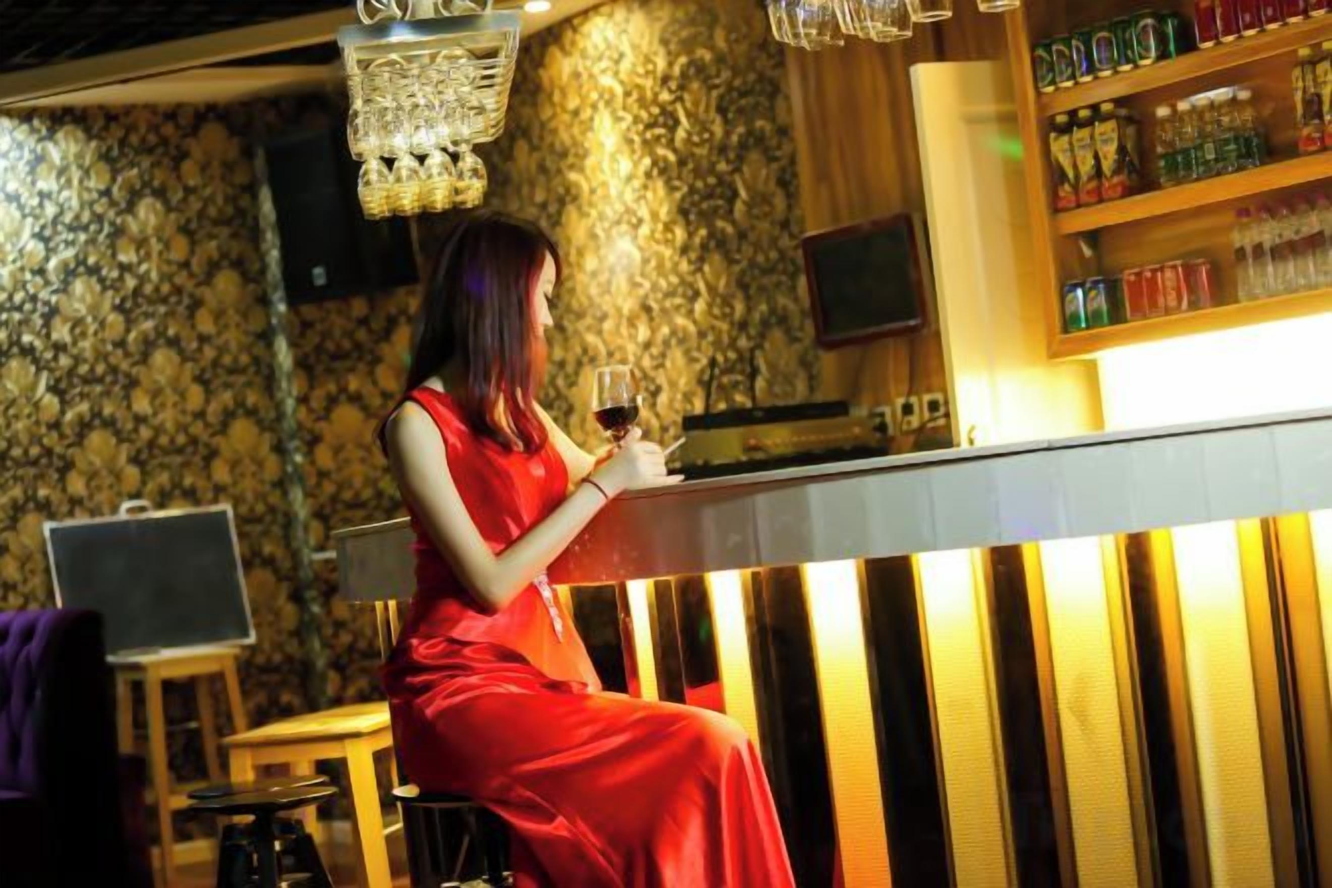 Una mujer con un vestido rojo sentada en un bar, con una bebida, un portátil sobre un taburete y un televisor en la pared.
