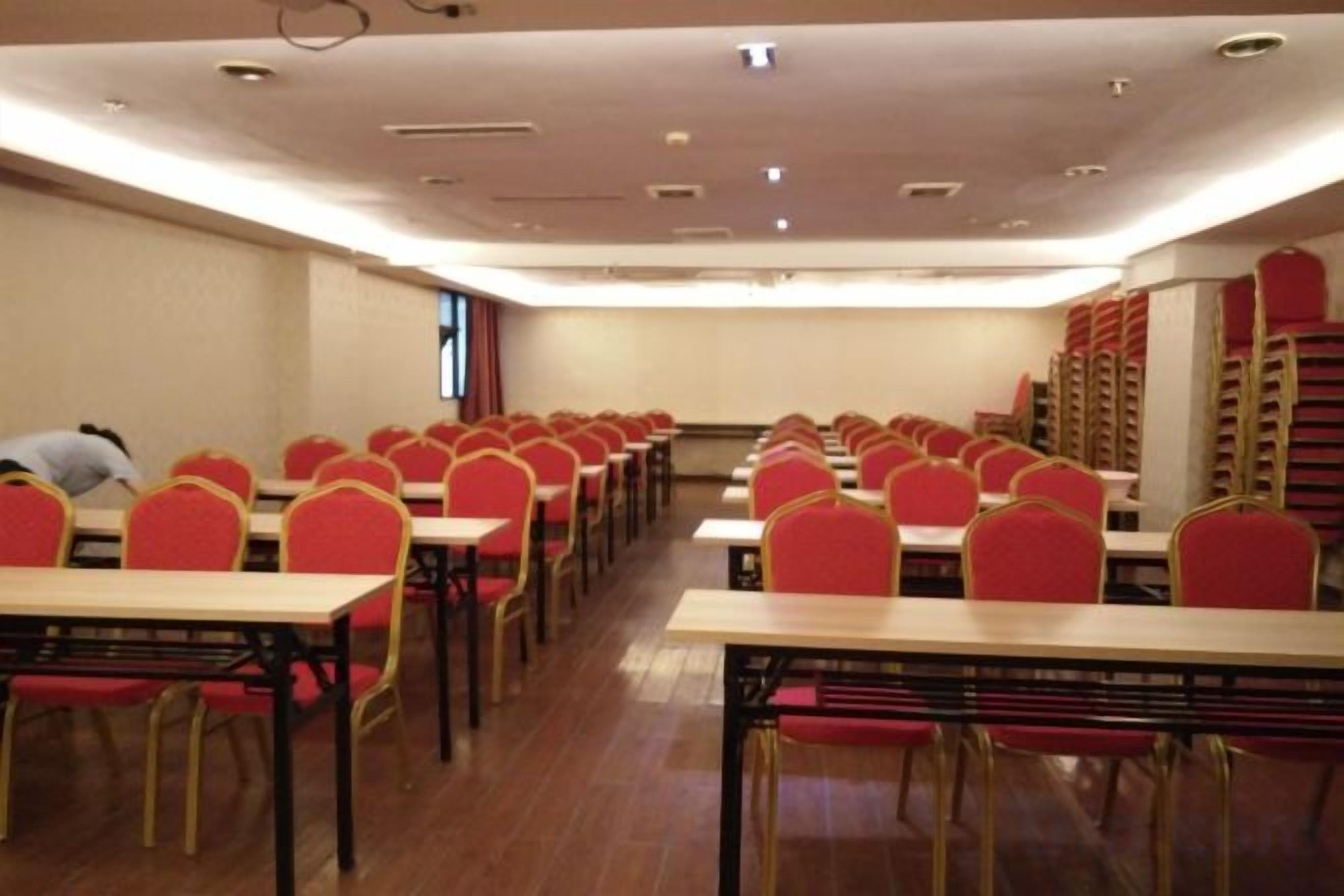 Uma sala de conferências com mesas e cadeiras dispostas em fileiras, uma tela de projetor e uma janela com cortinas.