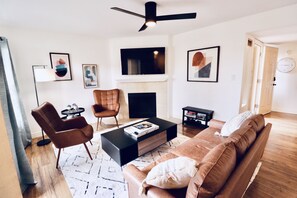 A modern living room with a brown leather sofa, a black coffee table, and two brown armchairs. A fireplace is centered on the wall, and there are framed artworks above it. - Mountain View Pickleball Place (Scottsdale)
