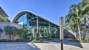 A modern building with large glass windows and a curved roof, surrounded by a landscaped area with palm trees and potted plants. - Modern Irvine Condo w/ Pool - 7 Mi to Beach! (Irvine)