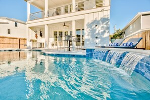 En swimmingpool med et vandfald, en overdækket terrasse med udendørs møbler, og en toetagers bygning med balkon.