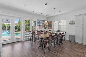 Un vaste espace dédié à la salle à manger, avec une grande table en bois, de nombreux tabourets de bar, un éclairage suspendu et une vue sur une piscine, visible à travers de grandes portes vitrées.