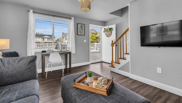 A modern living room with a grey sofa, a wooden coffee table with a tray, a chandelier, a TV mounted on the wall, and a staircase leading to a balcony. - Barnegat Bay Cottage: A 'MyShoreCottage' Property (South Seaside Park)