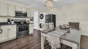 A modern kitchen with white cabinetry, stainless steel appliances, and a dining area with a glass table and chairs. - Barnegat Bay Cottage: A 'MyShoreCottage' Property (South Seaside Park)