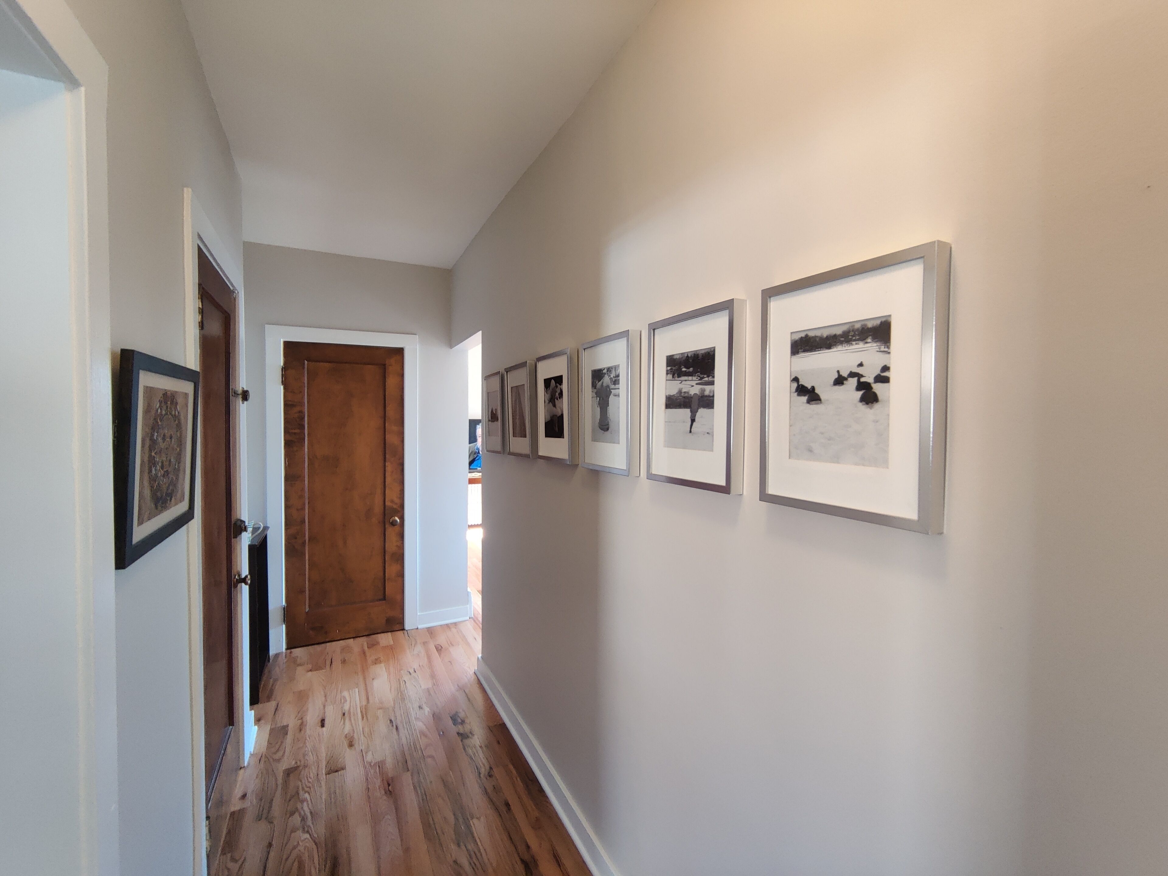 A hallway with framed photographs on the wall, wooden flooring, and a wooden door at the end.
