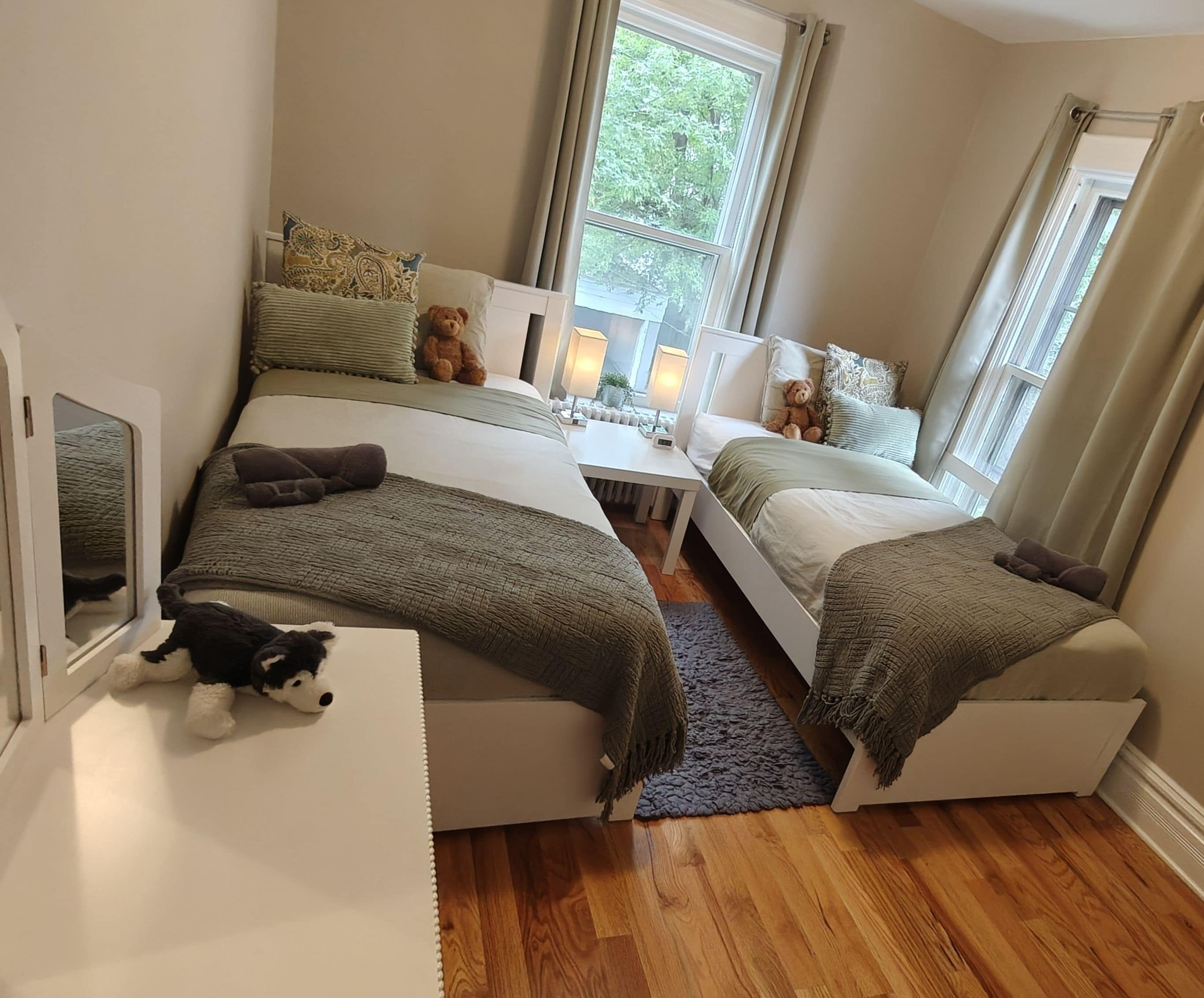 A cozy bedroom with two beds, a bedside table with lamps, and a teddy bear on the floor.