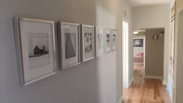 A hallway with framed pictures on the wall, leading to a bedroom with a bed and a painting on the wall.