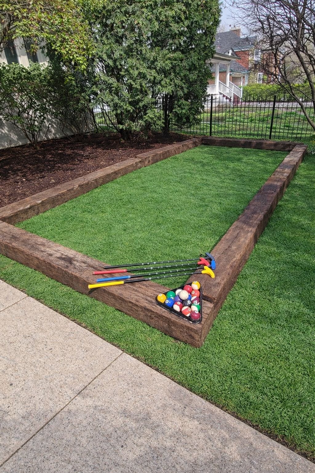 A pool table with balls and cues in a grassy area with a wooden border.