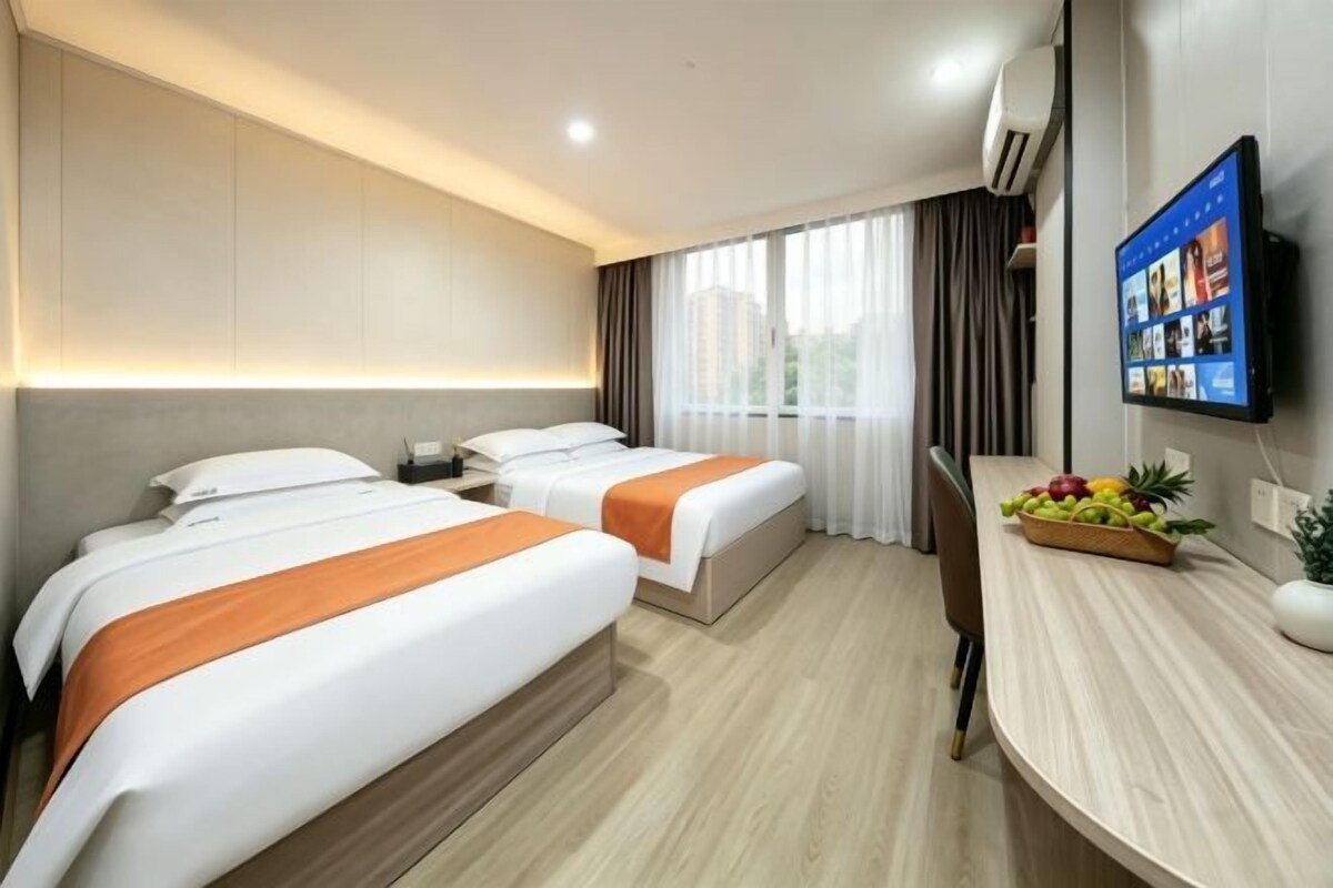 A hotel room with two beds, a desk with a fruit bowl, a flat-screen TV, and a window with curtains.