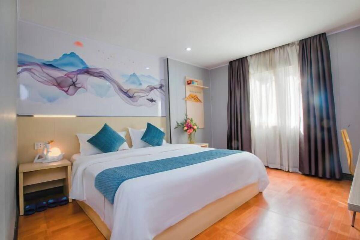 A hotel room with a bed, a nightstand, a wall-mounted artwork, a window with curtains, and a wooden floor.