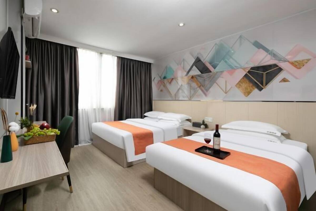 A hotel room with two beds, a desk with fruit, a bottle of wine, and a geometric wall design.