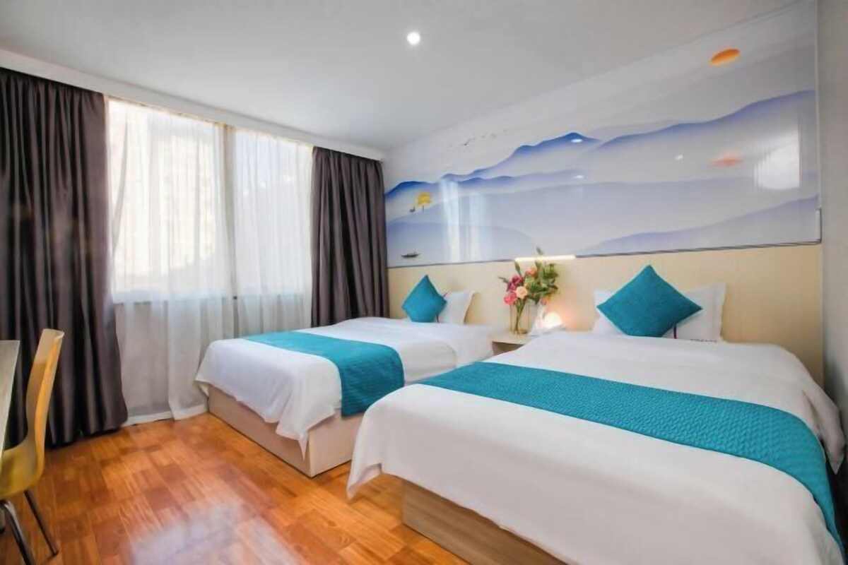 A hotel room with two beds, wooden flooring, a desk, and a large window with curtains.