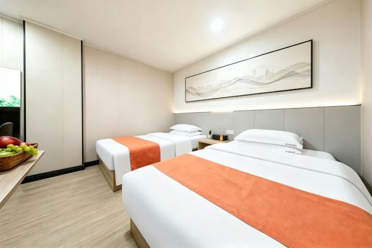A hotel room with two beds, a desk with fruit, and a wall-mounted artwork.