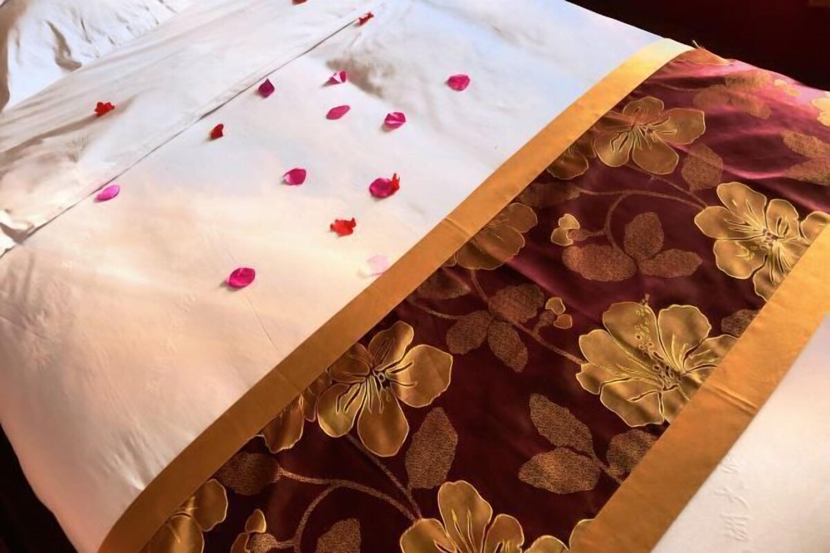 A bed with a white and maroon bedspread, adorned with gold floral patterns and pink rose petals.