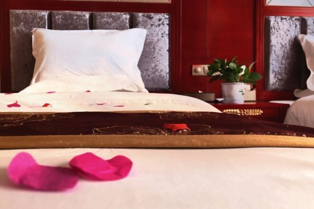 A bed with white linens and rose petals, a potted plant, and a red wall.