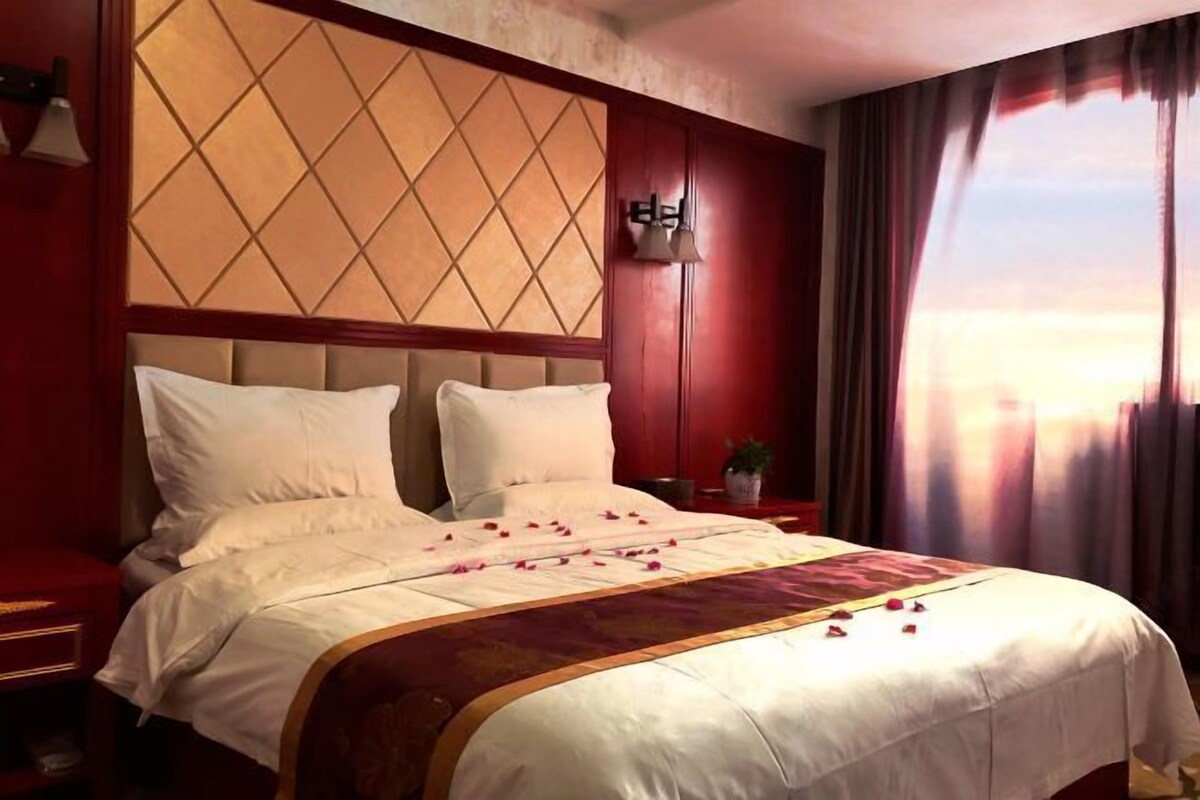 A hotel room with a large bed, a headboard with a diamond pattern, a nightstand with a lamp, and a window with curtains.