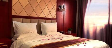 A hotel room with a large bed, a headboard with a diamond pattern, a nightstand with a lamp, and a window with curtains.