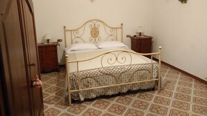 Un dormitorio con estructura de cama metálica, ropa de cama blanca, dos mesitas de noche con lámparas, un armario de madera y baldosas de suelo estampadas.