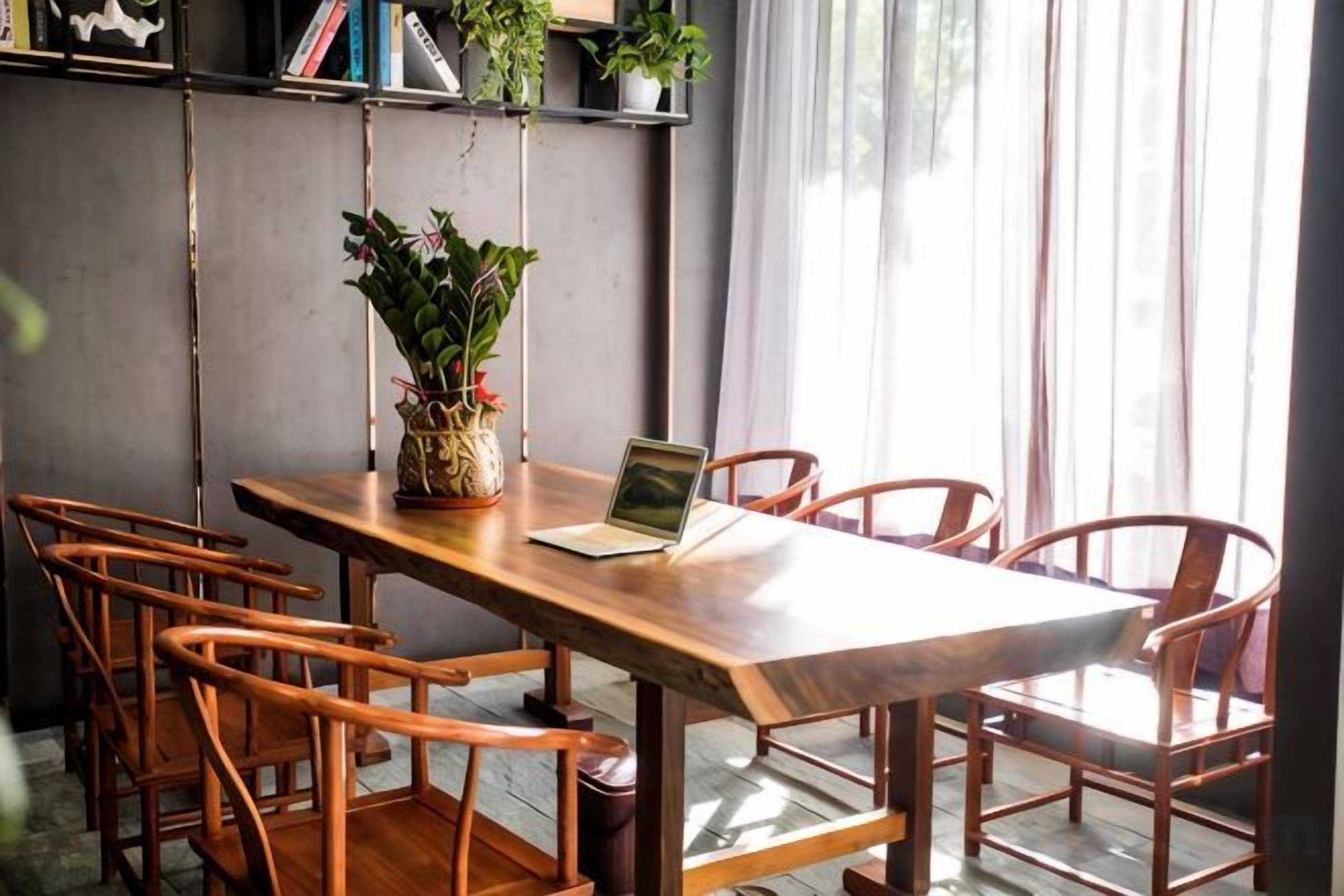 A wooden table with a laptop, surrounded by chairs, in a room with a large window and curtains.