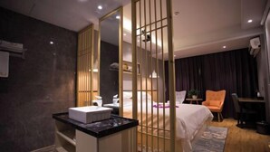 Een moderne hotelkamer met een bed, een bureau, een stoel, een badkamer met een wastafel en spiegel, en een decoratieve scheidingswand.