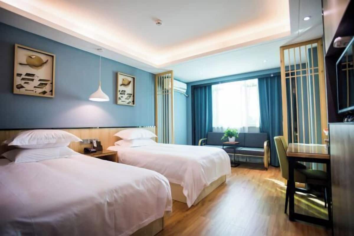 Ein Hotelzimmer mit zwei Betten, einem Schreibtisch, einem Stuhl, einem Fenster mit Vorhängen und Kunstwerken an den Wänden.