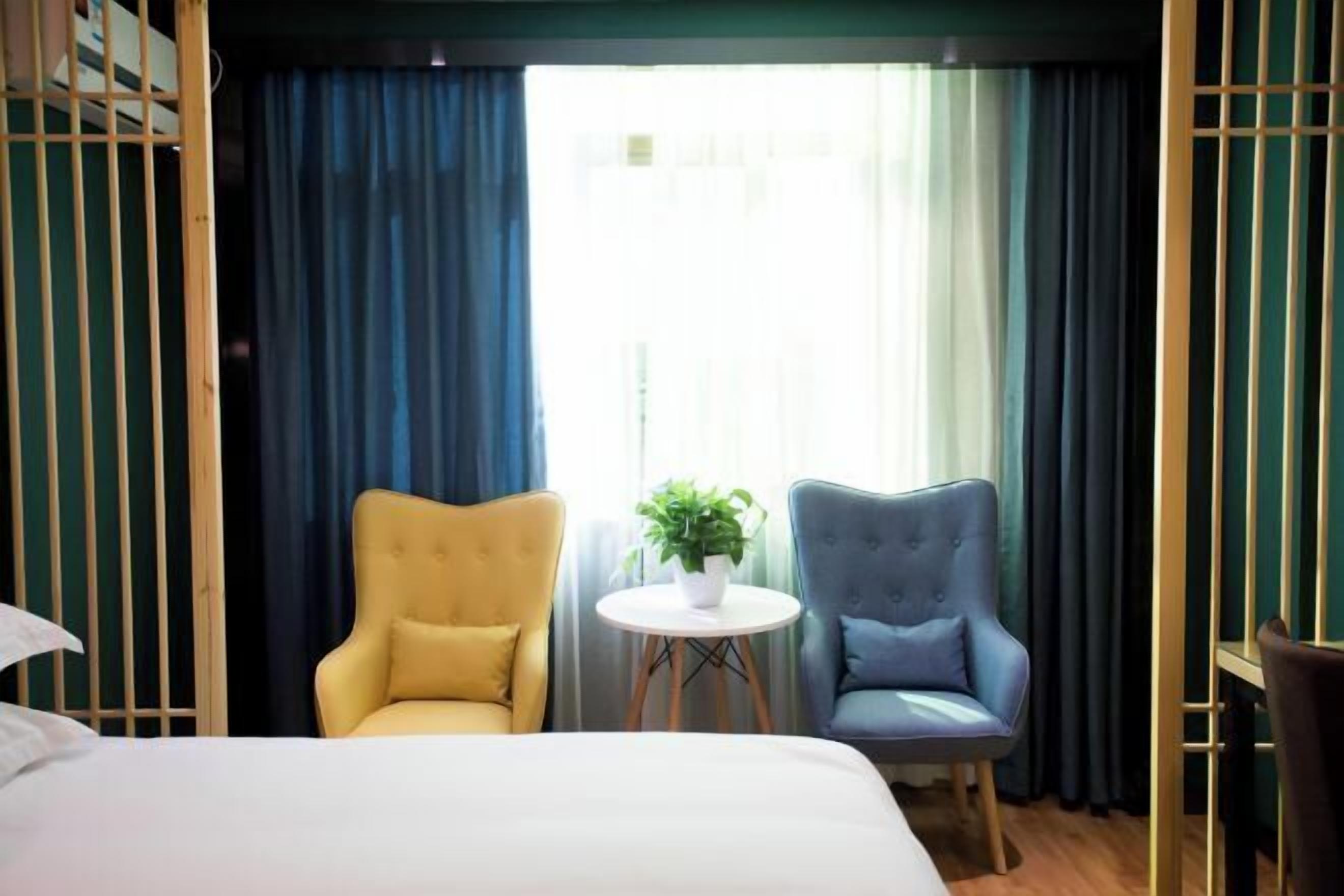 Une chambre d’hôtel avec un lit, deux fauteuils, une petite table ronde ornée d’une plante, et une vue sur l’extérieur à travers des rideaux transparents.
