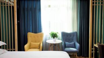 Une chambre d’hôtel avec un lit, deux fauteuils, une petite table ronde ornée d’une plante, et une vue sur l’extérieur à travers des rideaux transparents.