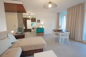 Una sala moderna con sofá, una mesa de comedor con sillas, zona de cocina con lavadora y un amplio ventanal con cortinas.