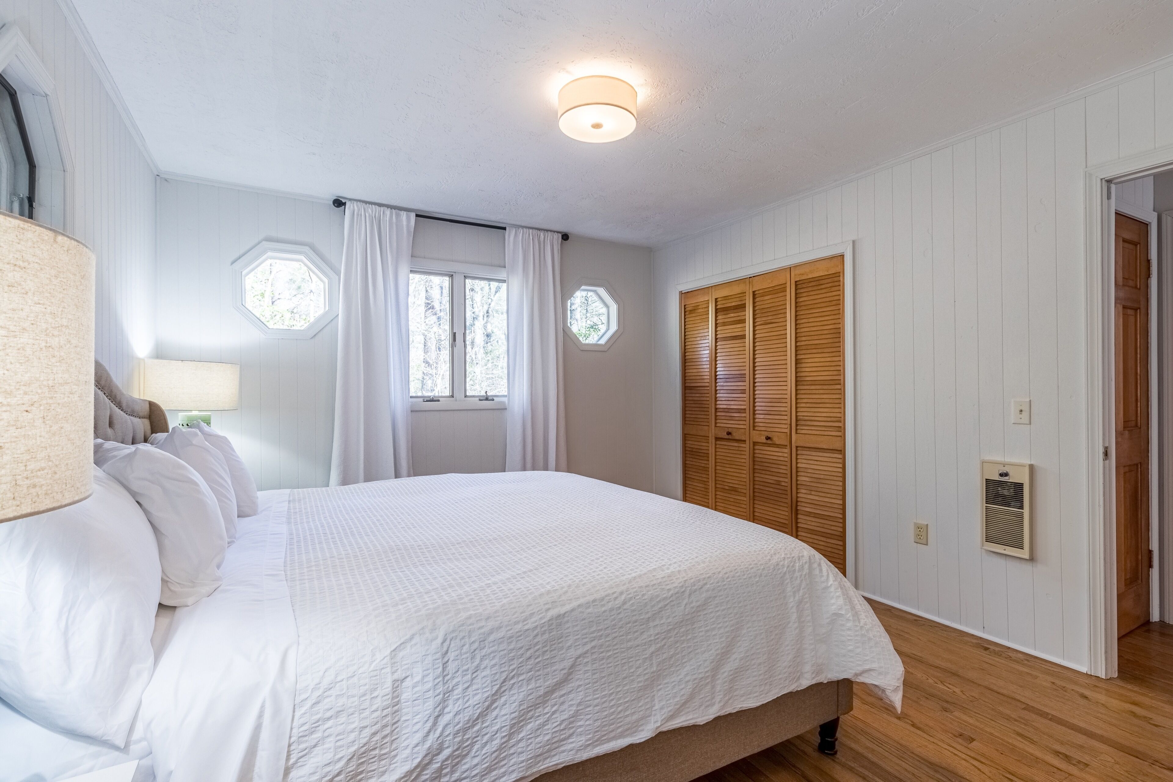 Une chambre à coucher avec un grand lit, une fenêtre avec des rideaux, une armoire en bois et un luminaire au plafond.
